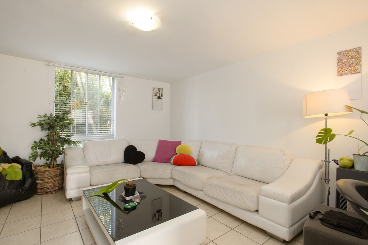 A bright living room featuring neutral tiled flooring, a large window with venetian blinds, and a central ceiling light fixture.