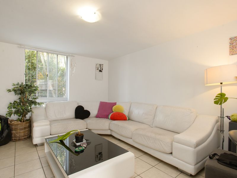 A bright living room featuring neutral tiled flooring, a large window with venetian blinds, and a central ceiling light fixture.