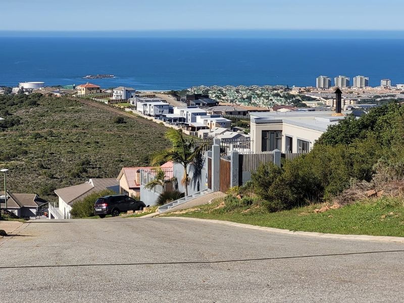 A street-level view looking down a residential road towards a coastal landscape with ocean views in the distance.