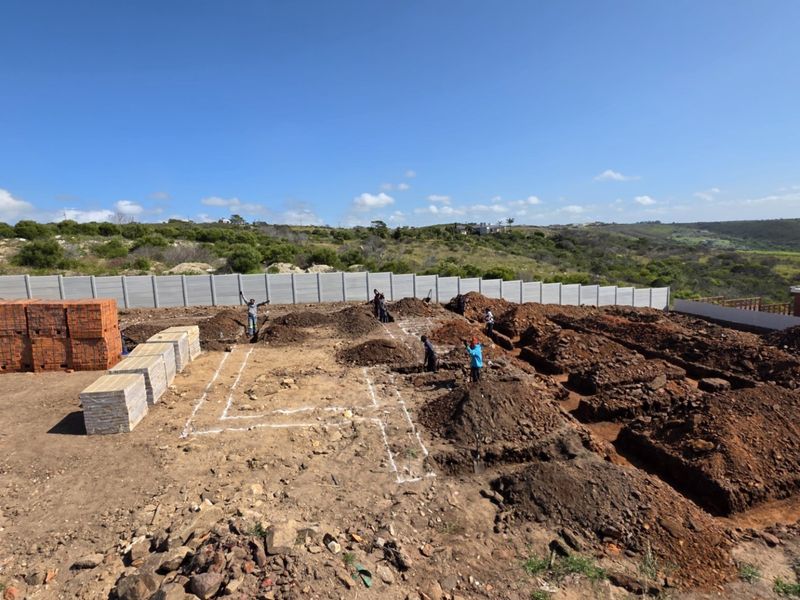 A construction site showing excavated foundation trenches and stacked building materials, enclosed by a precast concrete boundary wall.