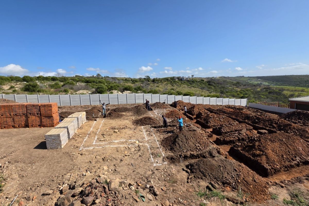 A construction site showing excavated foundation trenches and stacked building materials, enclosed by a precast concrete boundary wall.