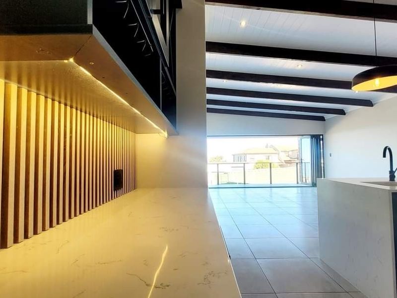 Modern open-plan kitchen and living area featuring stone countertops, slatted timber accents, exposed ceiling beams, and large bi-fold doors leading to an outdoor space.