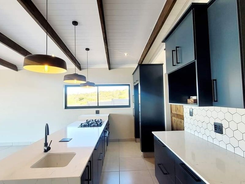 Modern kitchen featuring a central island with gas hob, stone countertops, dark cabinetry, hexagonal tile backsplash, and exposed ceiling beams.