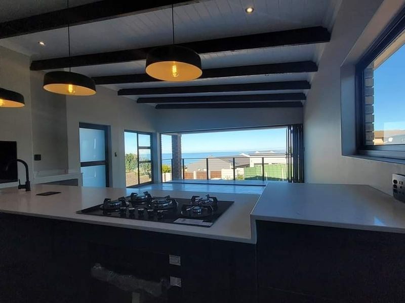 Modern open-plan kitchen and living area featuring exposed ceiling beams, stone countertops, a built-in gas hob, and expansive sliding doors with ocean views.