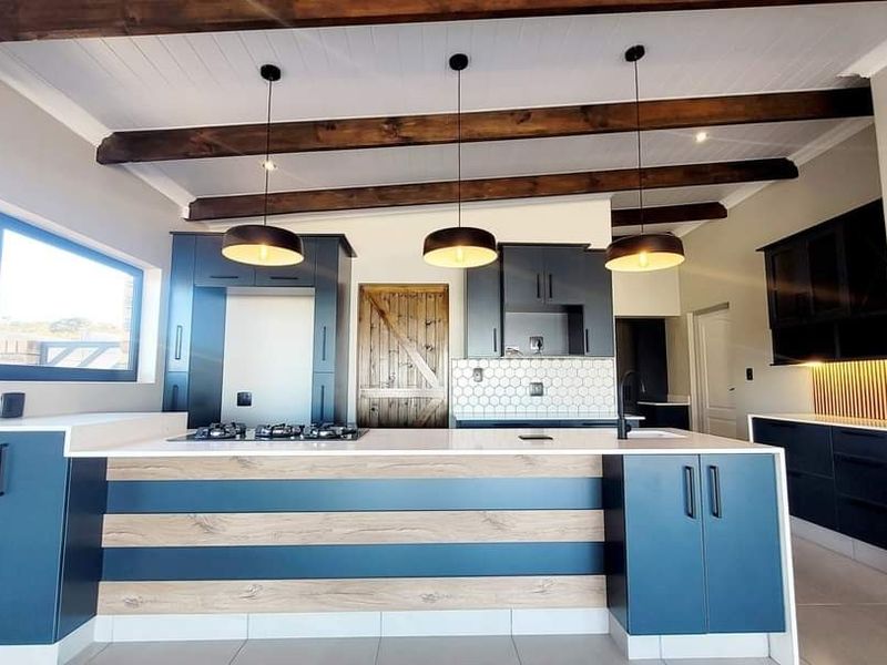 Modern kitchen featuring a large island with gas cooktop, dark blue and wood-tone cabinetry, stone countertops, and exposed timber ceiling beams.