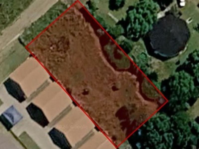An aerial view of a vacant residential plot of land, outlined in red, situated within a developed neighborhood.