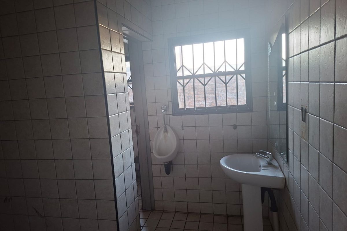 A functional commercial bathroom featuring a pedestal basin, wall-mounted urinal, and fully tiled surfaces with security bars on the window.