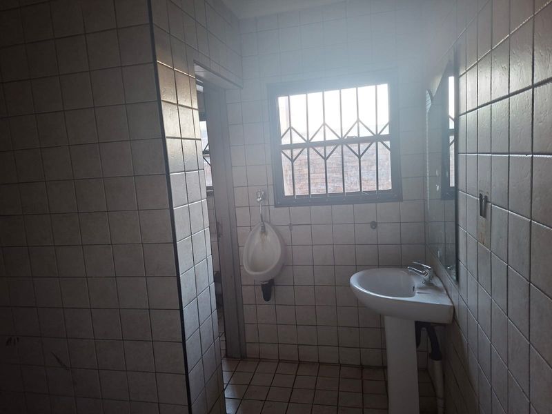 A functional commercial bathroom featuring a pedestal basin, wall-mounted urinal, and fully tiled surfaces with security bars on the window.