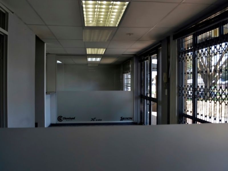 An empty office space featuring a suspended ceiling with fluorescent lighting, glass partitioning, and a security gate at the entrance.