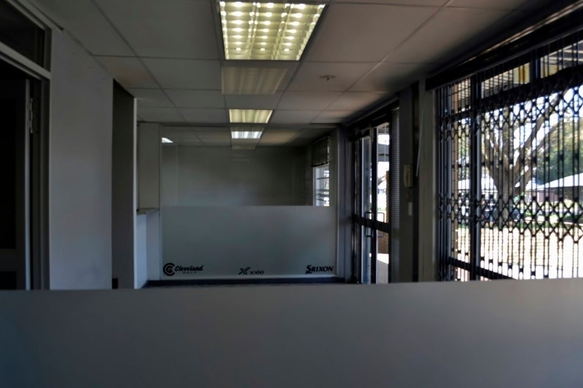 An empty office space featuring a suspended ceiling with fluorescent lighting, glass partitioning, and a security gate at the entrance.