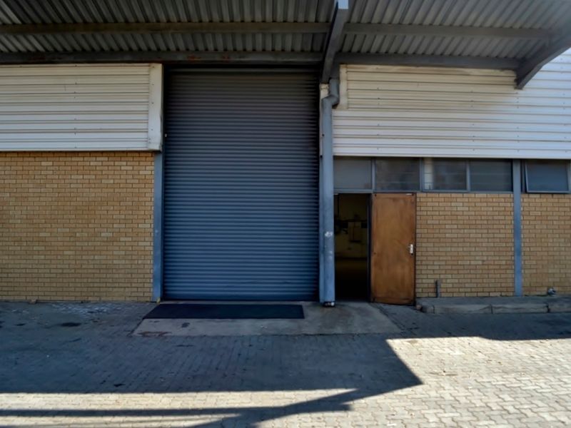 Industrial warehouse exterior featuring a large roller shutter door, pedestrian access door, brick facade, and paved loading area under a metal roof canopy.