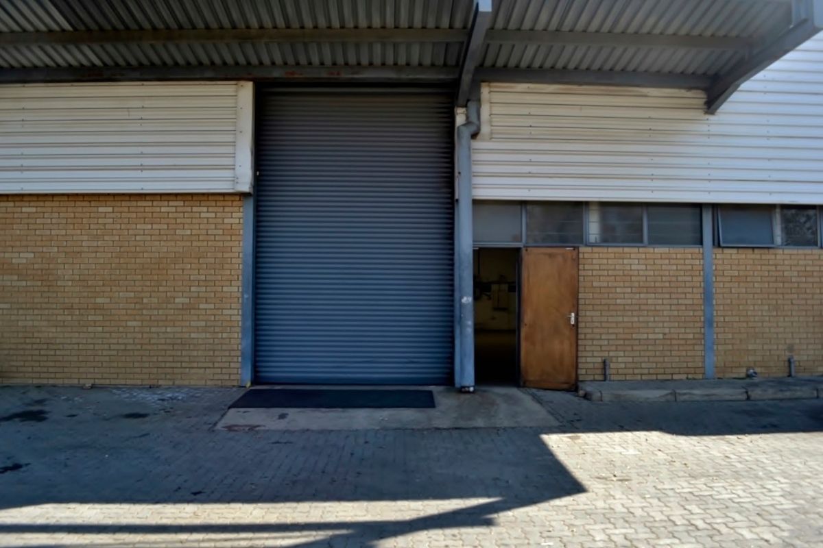 Industrial warehouse exterior featuring a large roller shutter door, pedestrian access door, brick facade, and paved loading area under a metal roof canopy.