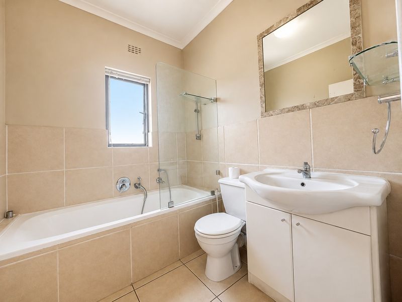 A clean, neutral-toned bathroom featuring a built-in bathtub with a glass shower screen, a vanity unit with a basin, and a wall-mounted mirror.