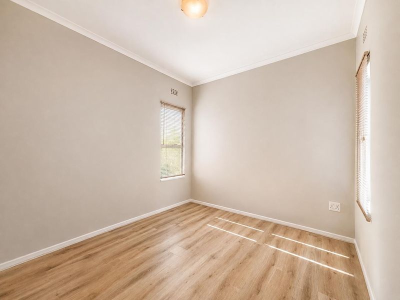 A bright, empty bedroom featuring modern laminate flooring, neutral walls, and venetian blinds, offering a clean and versatile space.