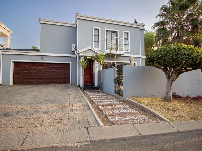 Front elevation of a two-story house featuring a paved driveway, double garage, balcony, and a gated pedestrian entrance with a landscaped walkway.