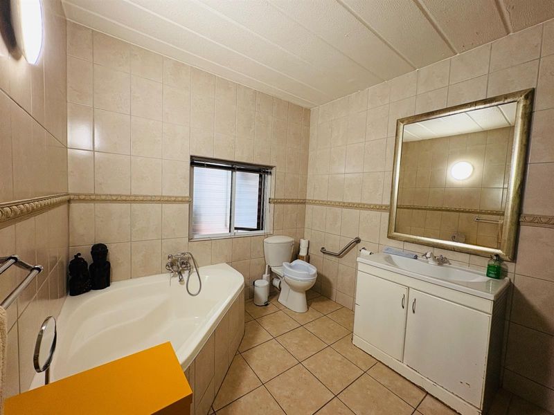 A functional bathroom featuring a corner bathtub, vanity cabinet with basin, toilet, and wall-mounted grab rail, finished with neutral wall and floor tiles.