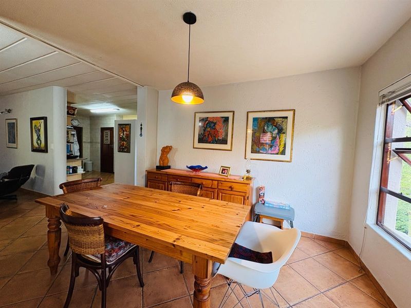 A dining room featuring terracotta floor tiles, a central pendant light, and natural light from a side window, connecting to an adjacent kitchen area.