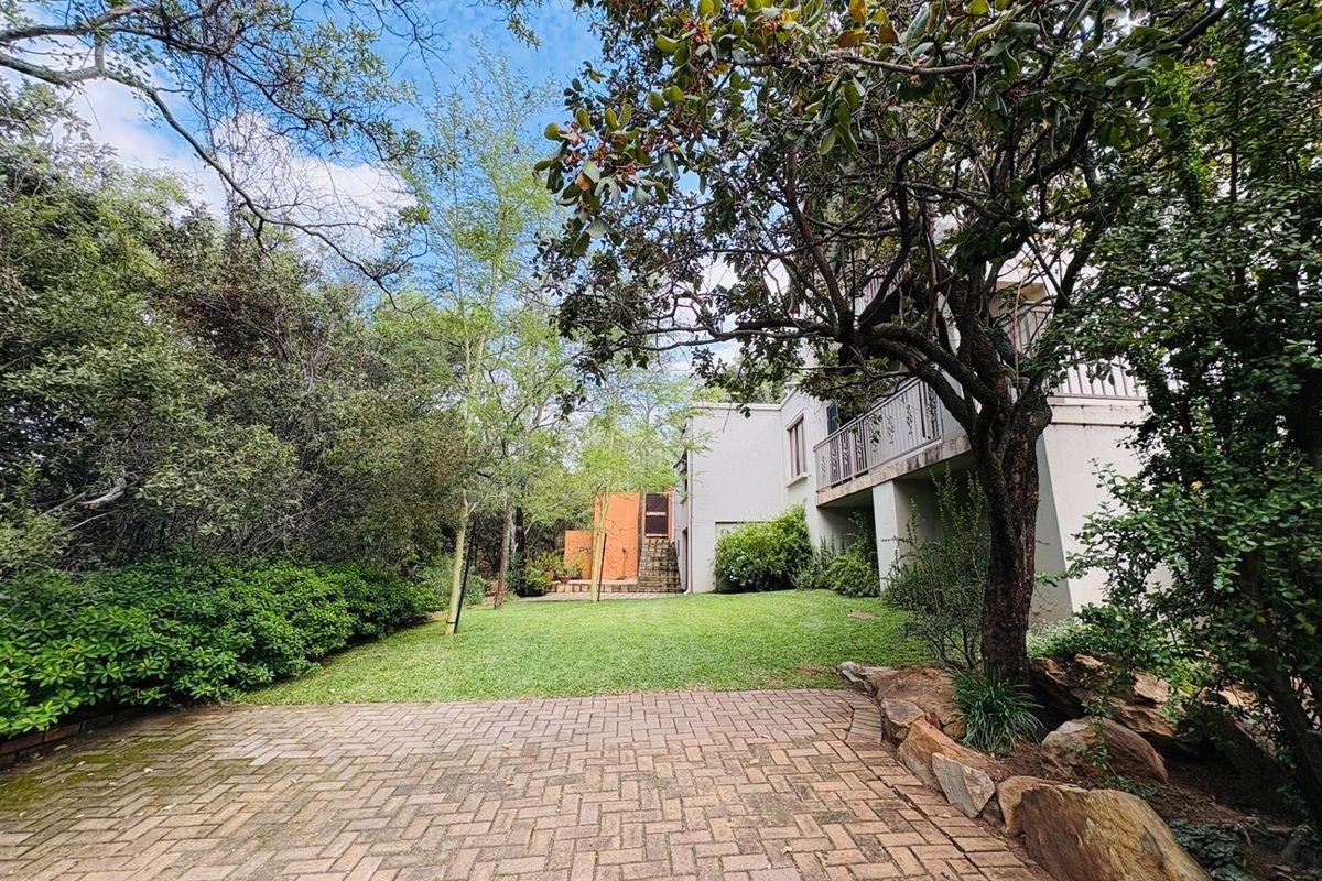 A spacious rear garden featuring a paved patio area, lush lawn, mature trees, and a multi-level building facade with a balcony.