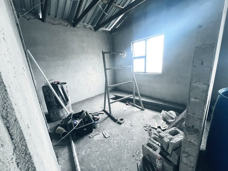 An unfinished bedroom space featuring exposed roof trusses, plastered walls, and a metal window frame, currently in a shell construction state.