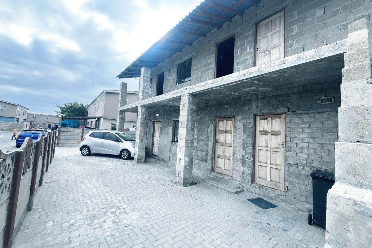 Two-story residential building featuring unfinished concrete block construction, multiple timber entry doors, and a paved driveway area.