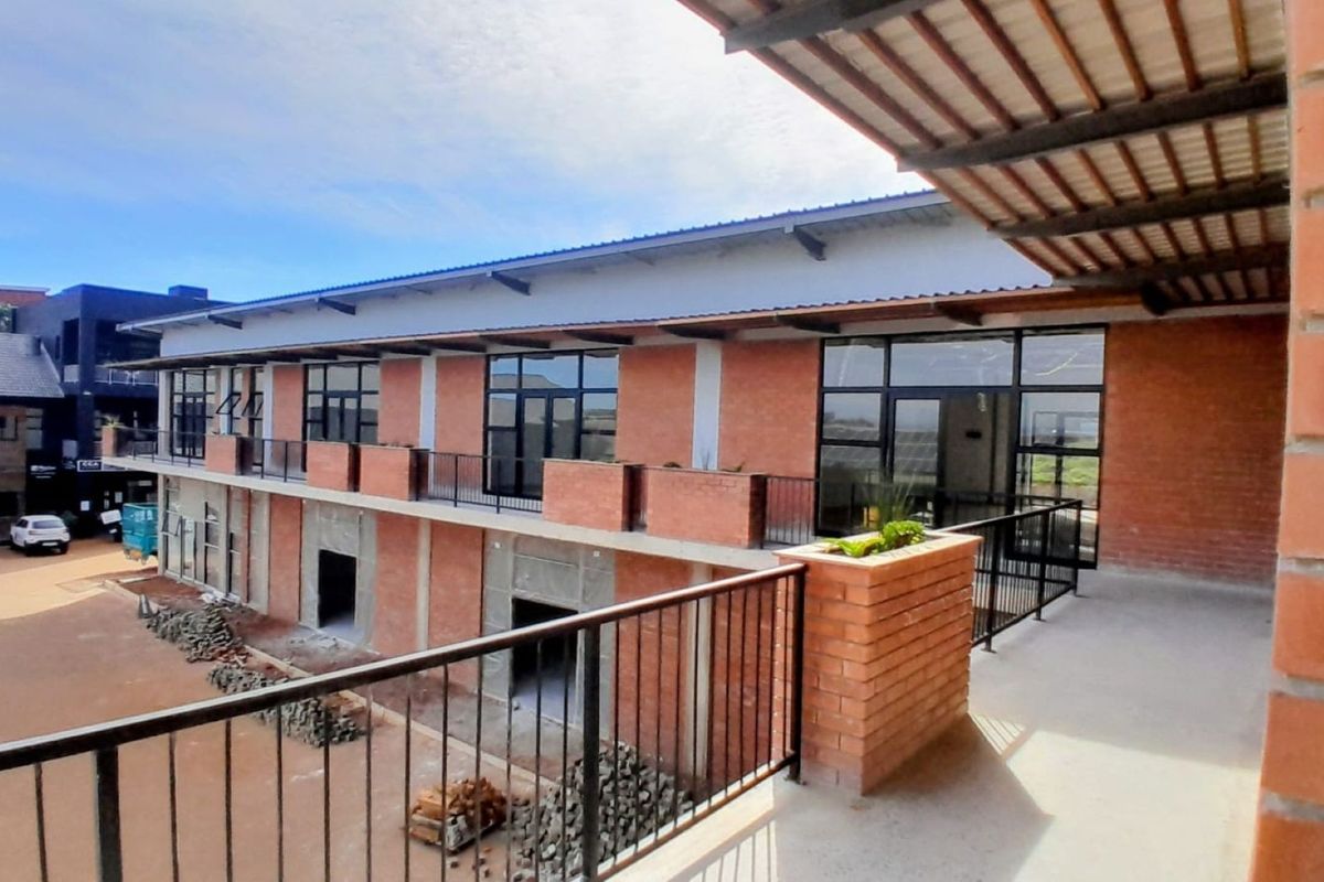 An exterior view of a modern office building featuring exposed brickwork, a covered balcony with metal railings, and large glass windows.