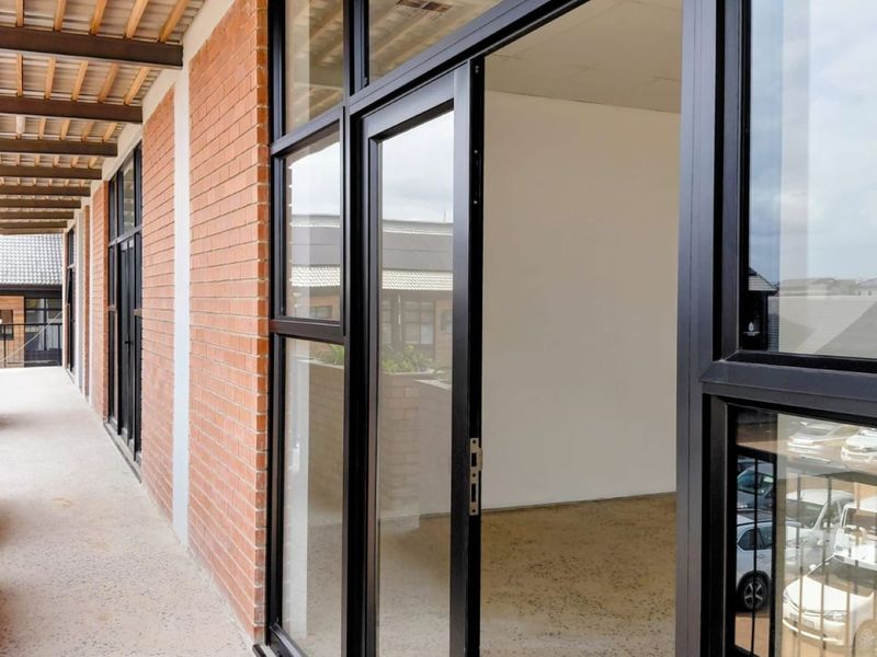 A modern covered balcony featuring exposed brick walls, timber rafters, and black aluminium framed glass doors leading into an office space.