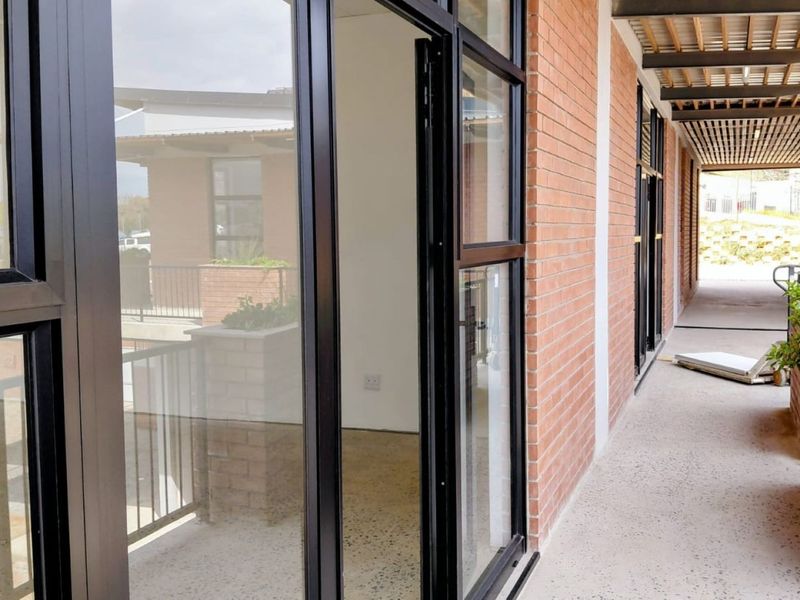 A modern covered balcony featuring exposed brick walls, sleek aluminium-framed glass doors, and a timber slat ceiling, overlooking the office complex exterior.