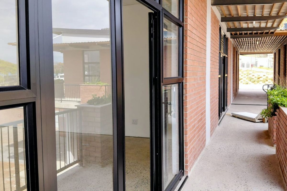 A modern covered balcony featuring exposed brick walls, sleek aluminium-framed glass doors, and a timber slat ceiling, overlooking the office complex exterior.