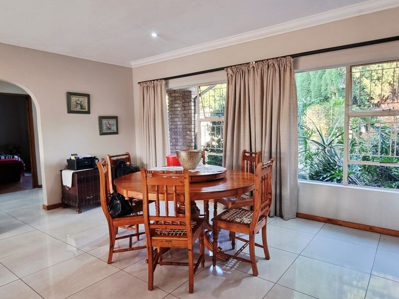 A bright dining room featuring large windows with garden views, neutral tiled flooring, and a classic arched doorway leading to adjacent living spaces.