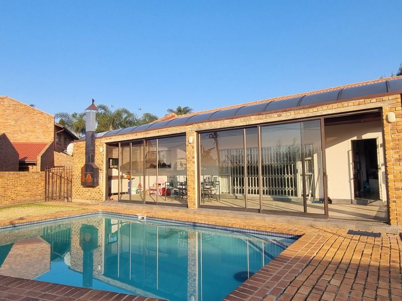 A well-maintained outdoor pool area featuring a brick-paved deck, a built-in outdoor fireplace, and a covered patio with sliding glass doors.