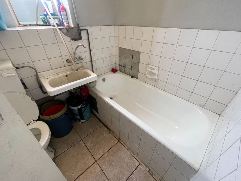 A compact bathroom featuring a bathtub, wall-mounted basin, and toilet, requiring significant renovation and tile repairs to restore functionality and aesthetic appeal.