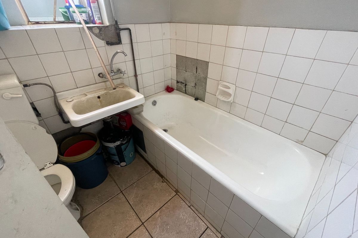 A compact bathroom featuring a bathtub, wall-mounted basin, and toilet, requiring significant renovation and tile repairs to restore functionality and aesthetic appeal.