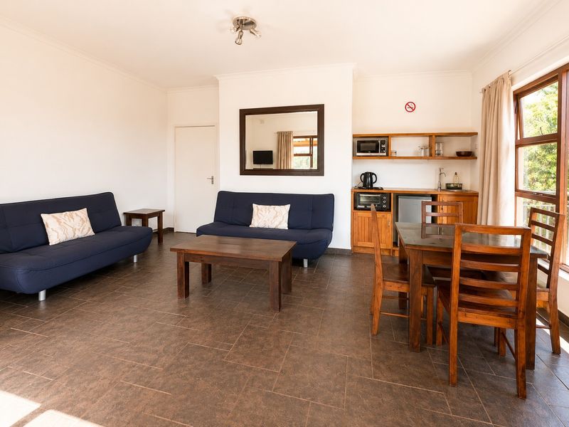 Open-plan living and dining area featuring tiled floors, a compact kitchenette with built-in cabinetry, and large windows providing natural light.