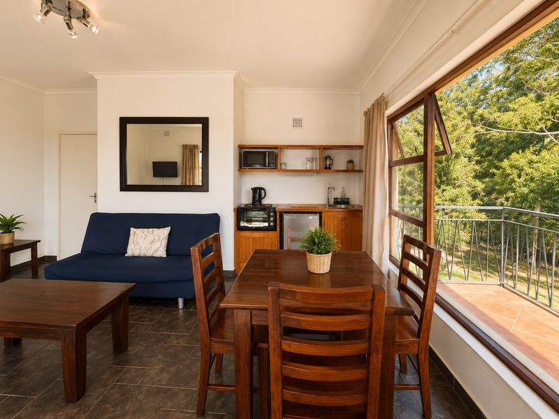 Open-plan living area featuring a compact kitchenette with wooden cabinetry, tiled floors, and large windows providing natural light and access to a balcony.