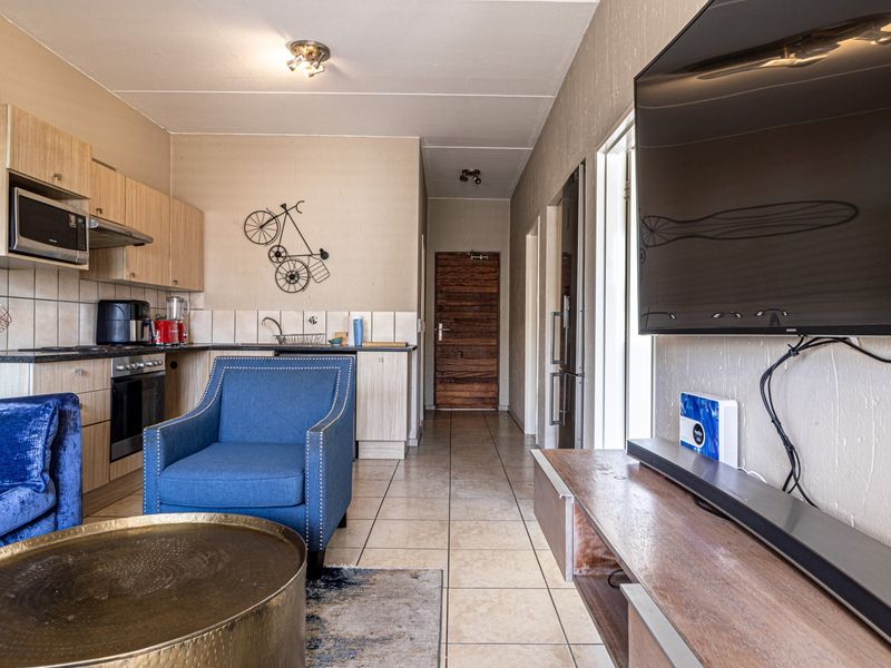 Open-plan living area featuring a compact kitchen with built-in cabinetry, integrated appliances, and tiled flooring, leading to a hallway with a solid wood door.