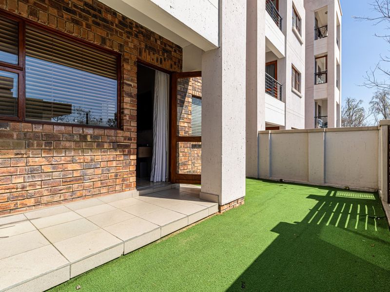 A private ground floor patio area featuring tiled flooring, an artificial grass lawn, and an exposed brick exterior wall with direct access to the apartment.