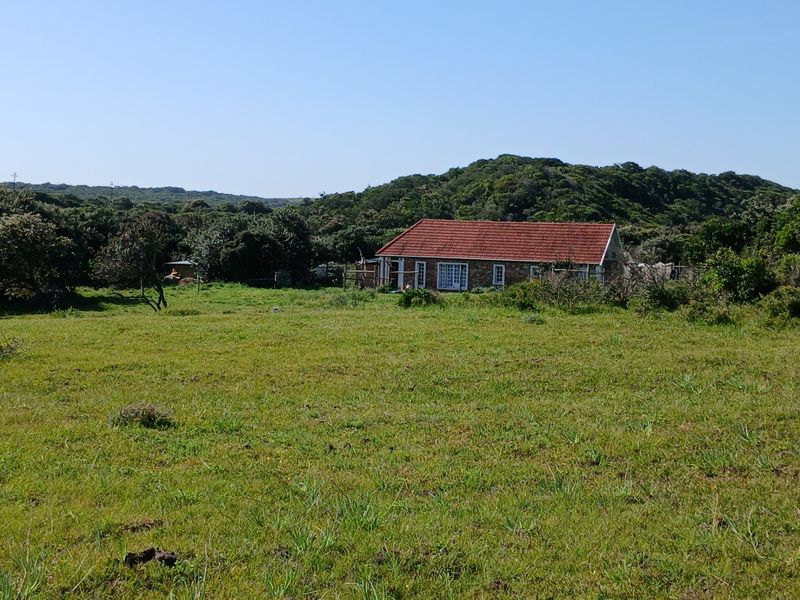 A view of the rear elevation of a stone-built cottage set within a large, open grassy field surrounded by natural vegetation.