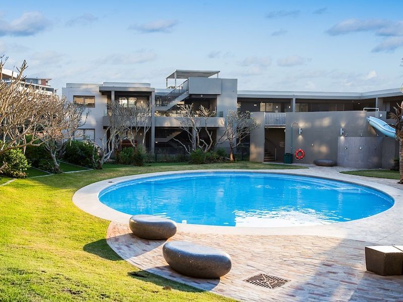 A well-maintained circular swimming pool area featuring a slide, paved surround, and landscaped garden set against a modern multi-level residential building.