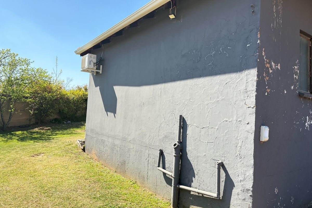 Rear exterior view of the property showing a garden area, external plumbing, and an air conditioning unit mounted on a wall with peeling paint.