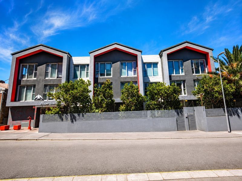 Front elevation of a modern apartment complex featuring a contemporary facade with red accents, secure perimeter fencing, and gated pedestrian access.
