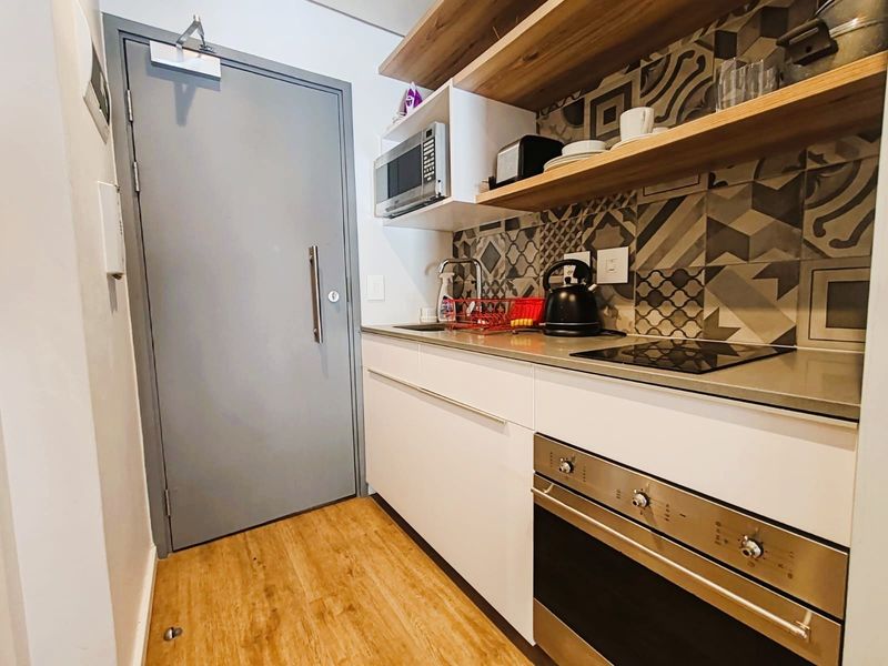 Compact modern kitchen featuring built-in oven, electric hob, microwave, patterned tile splashback, and floating timber shelving with durable laminate flooring.