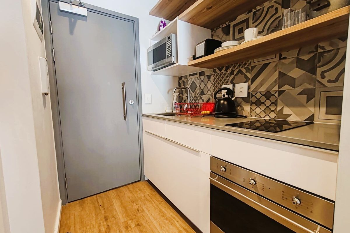 Compact modern kitchen featuring built-in oven, electric hob, microwave, patterned tile splashback, and floating timber shelving with durable laminate flooring.