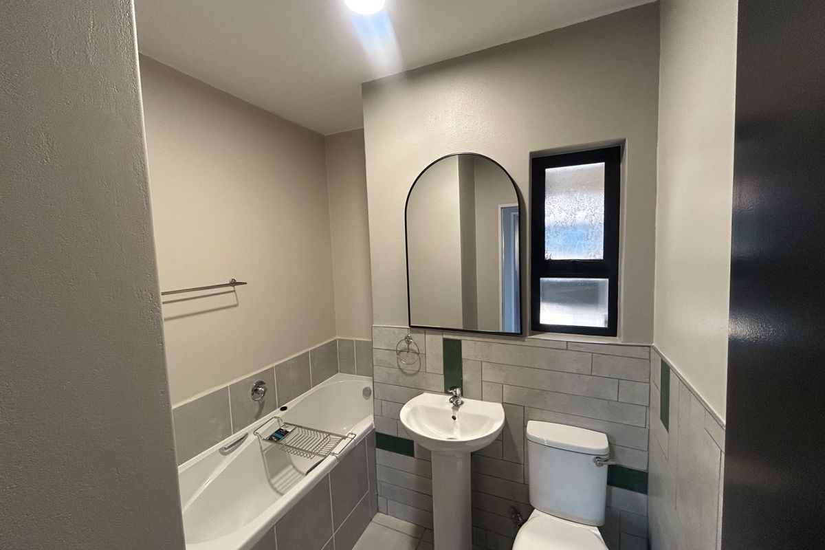 A clean, modern bathroom featuring a pedestal basin, bathtub, toilet, and stylish arched mirror with neutral wall finishes and tiled splashbacks.