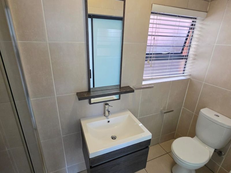 A modern, well-maintained bathroom featuring a vanity cabinet with basin, a wall-mounted mirror, a toilet, and a glass-enclosed shower with neutral wall tiling.