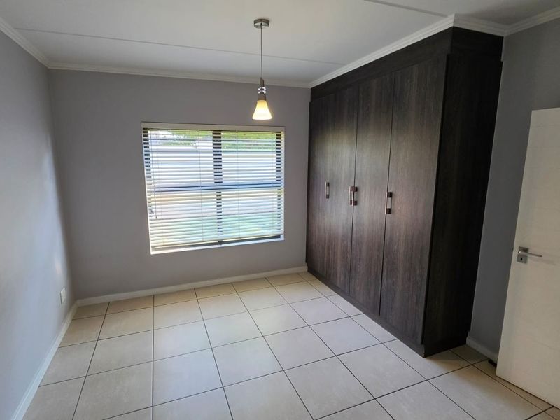 A clean, empty bedroom featuring neutral tiled flooring, built-in dark wood cupboards, and a window fitted with venetian blinds.