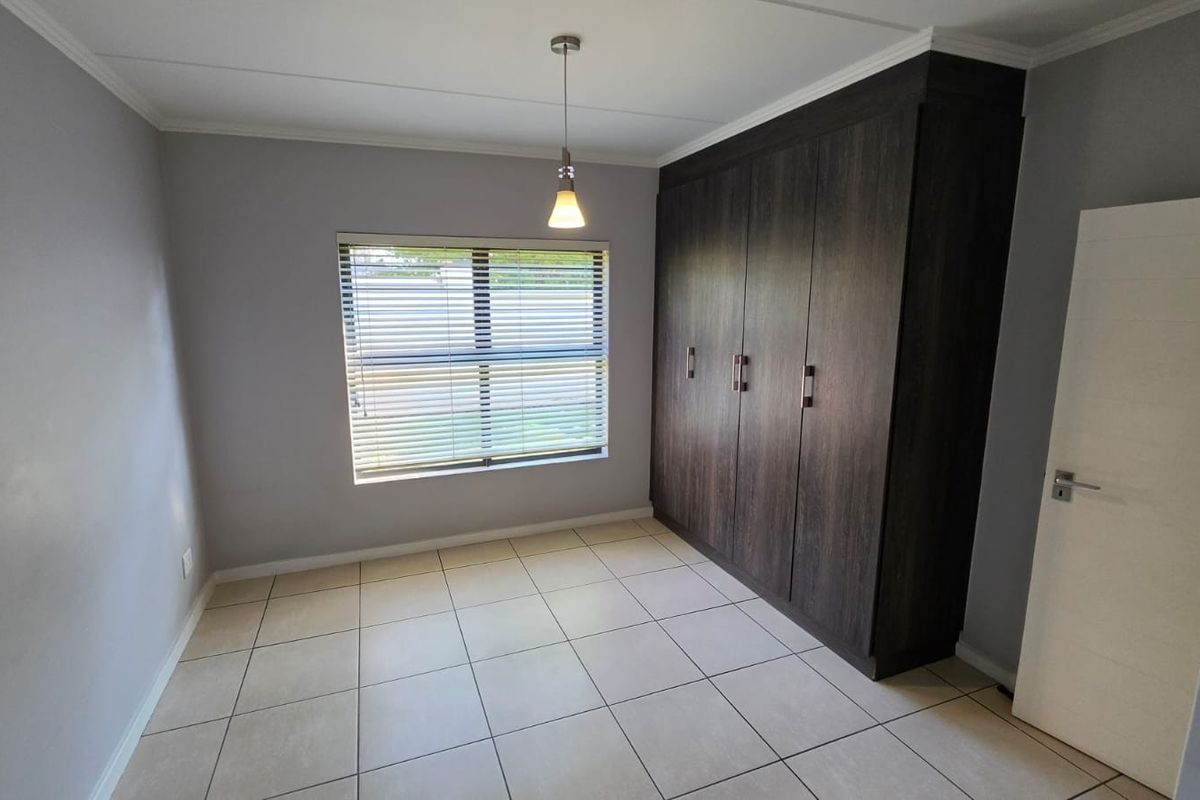 A clean, empty bedroom featuring neutral tiled flooring, built-in dark wood cupboards, and a window fitted with venetian blinds.