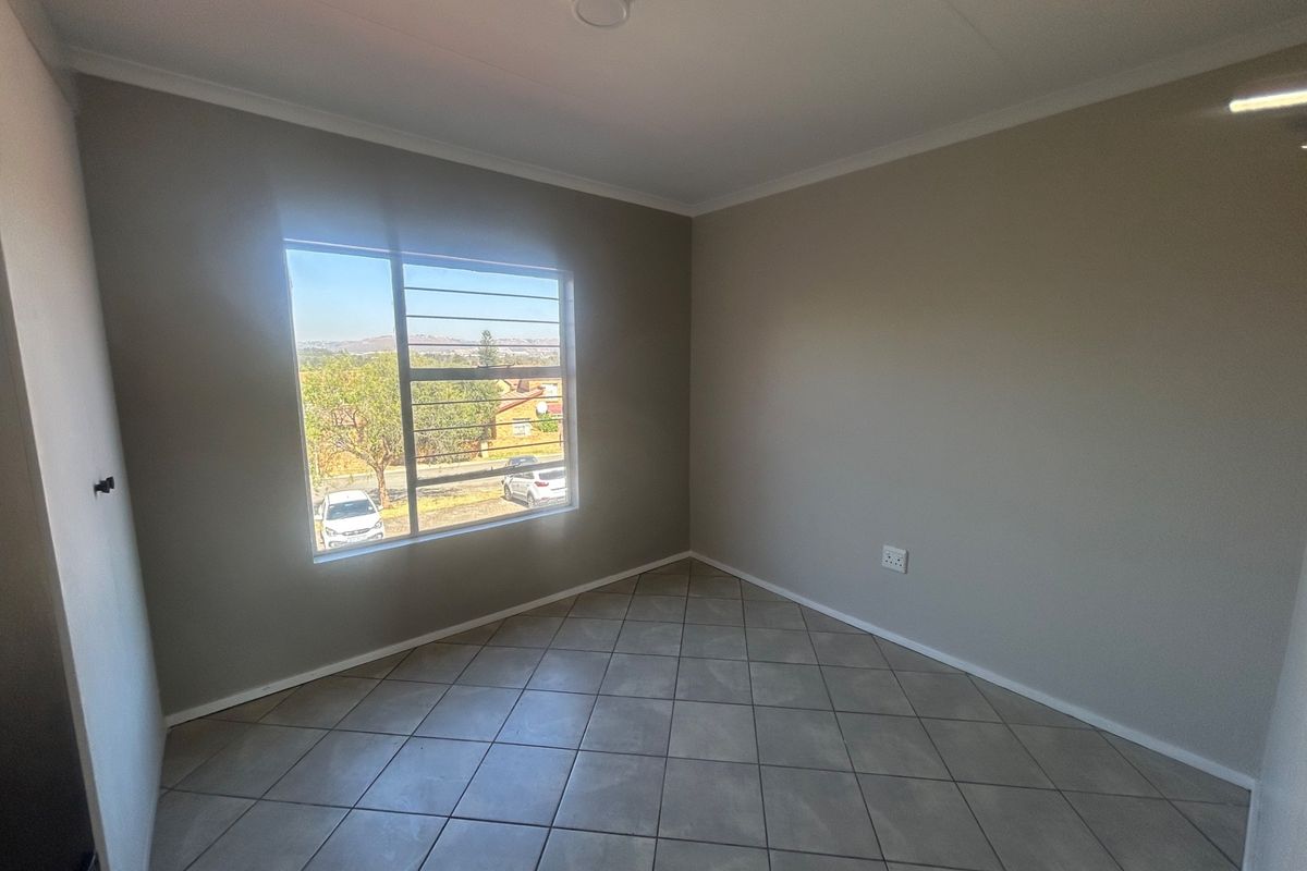 A bright, empty bedroom featuring neutral wall paint, tiled flooring, and a window with security bars offering an exterior view.