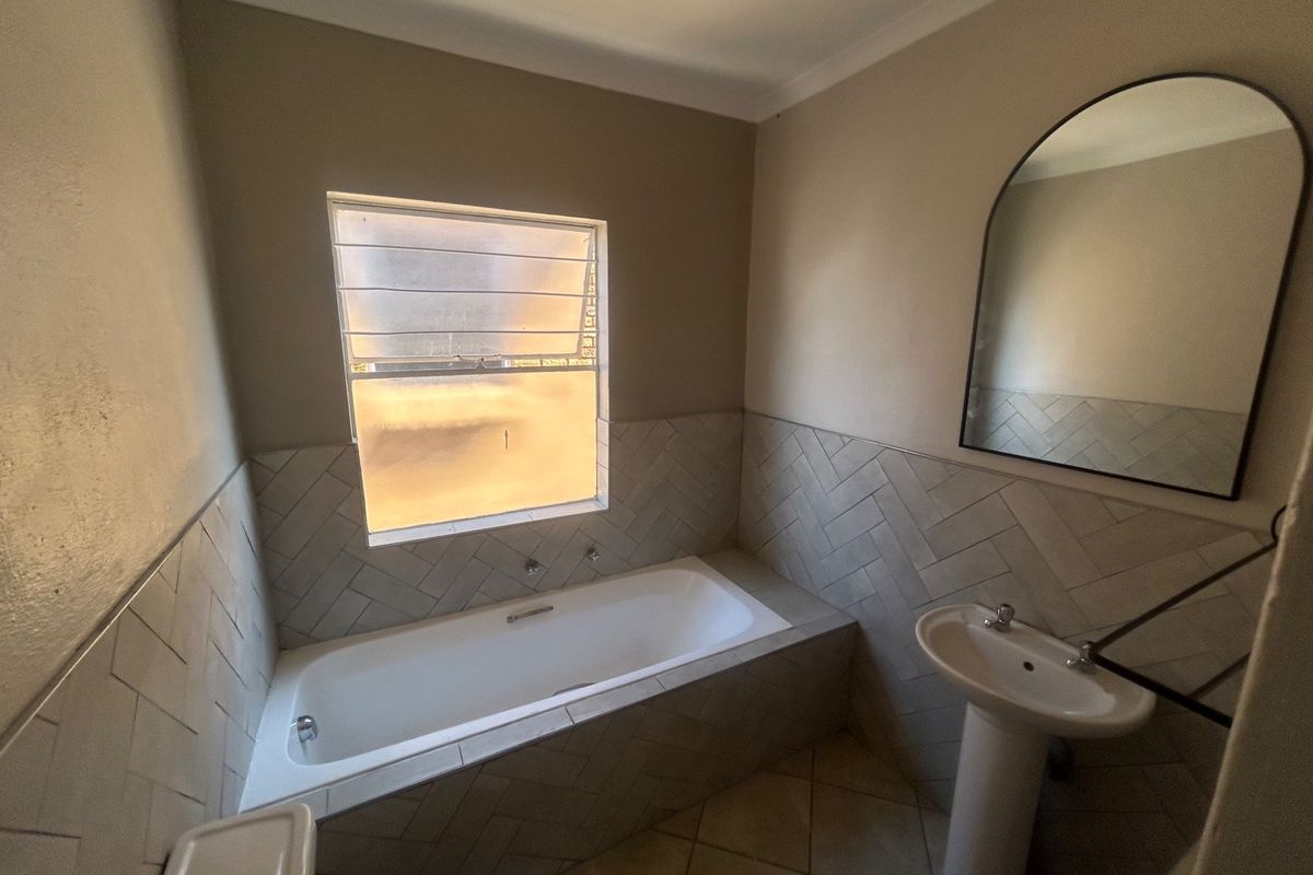A clean, functional bathroom featuring a built-in bathtub, pedestal basin, and stylish herringbone wall tiling with a large arched mirror.