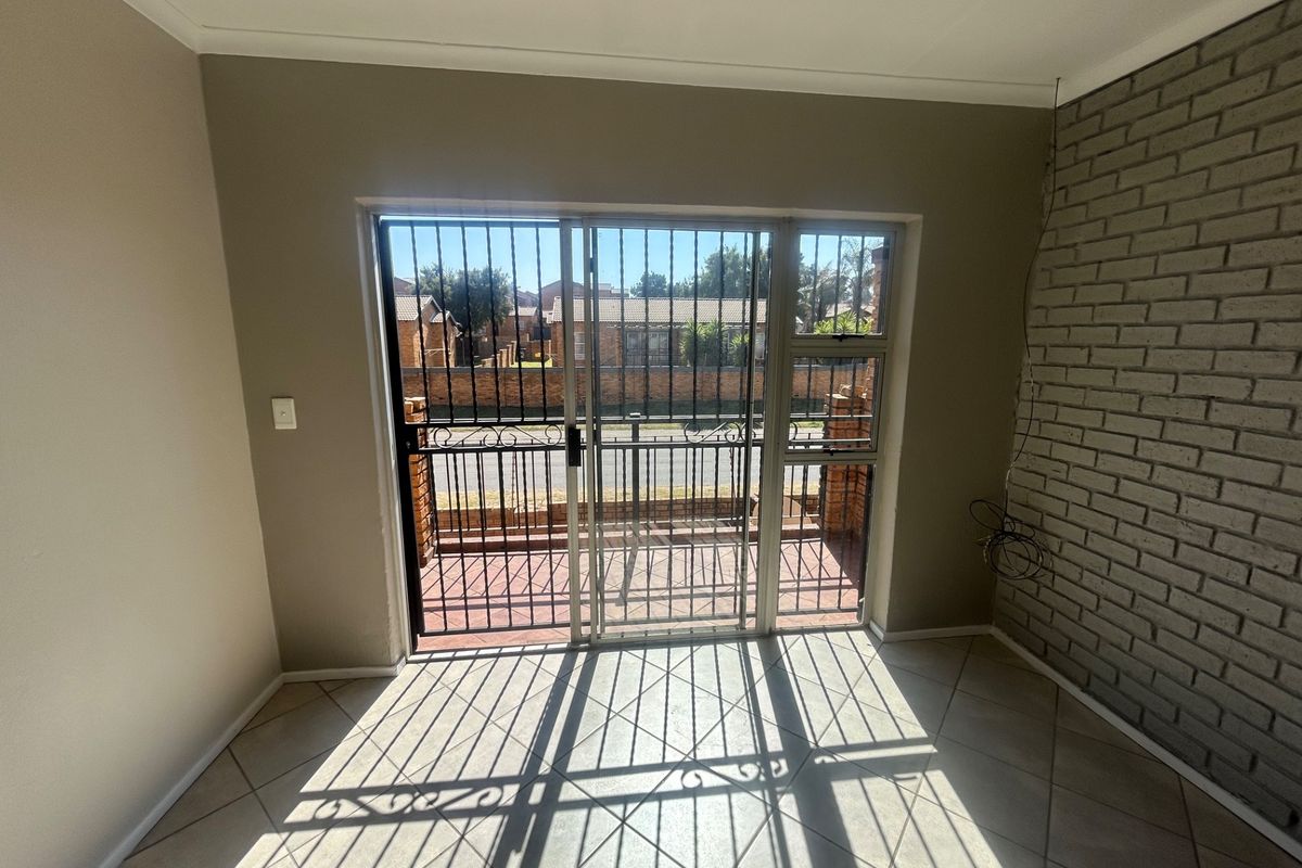 An empty living area featuring tiled floors, an exposed brick feature wall, and a sliding glass door with a security gate leading outside.