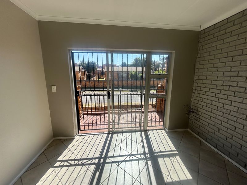 An empty living area featuring tiled floors, an exposed brick feature wall, and a sliding glass door with a security gate leading outside.